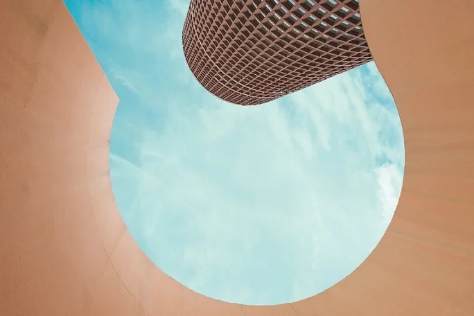 An abstract architectural scene shows a cylindrical tower framed by a large circular opening. The viewer looks upward from within a peach-colored structure toward the sky. The tower features a grid-like facade that contrasts with the smooth curved edges of the foreground. Soft blue sky and scattered clouds add a calm, minimal backdrop to the geometric composition.